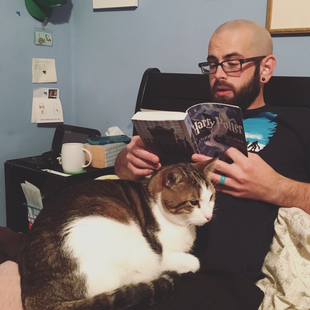 Joe is reading Harry Potter aloud while lying down. Nibbler, a tabby with a predominantly white face and belly, sits on top of him.