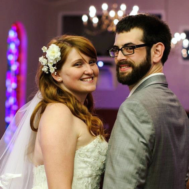 Joe and Sarah on their wedding day. They are both looking over their shoulders to smile for the camera. Sarah has a large white flower in her hair and Joe is in a dove grey suit. The chapel is backlit by purple light from stained glass windows.
