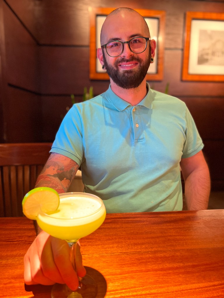 Joe is wearing a mint green polo and smiling as he holds a neon green cocktail on the table. The restaurant is backlit with orange light against modern wood walls.